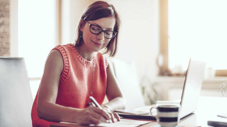 Woman sitting at computer, smiling and writing on a piece of paper